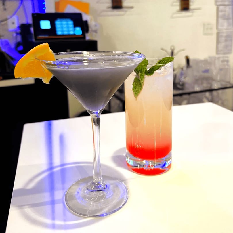 Two cocktails on a white table: a grey martini with an orange slice and a layered red and orange drink.