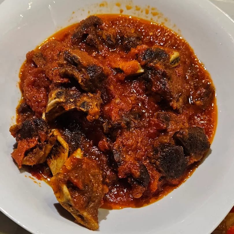 A close-up of African stewed goat meat in a rich, red tomato-based sauce, served in a white bowl.