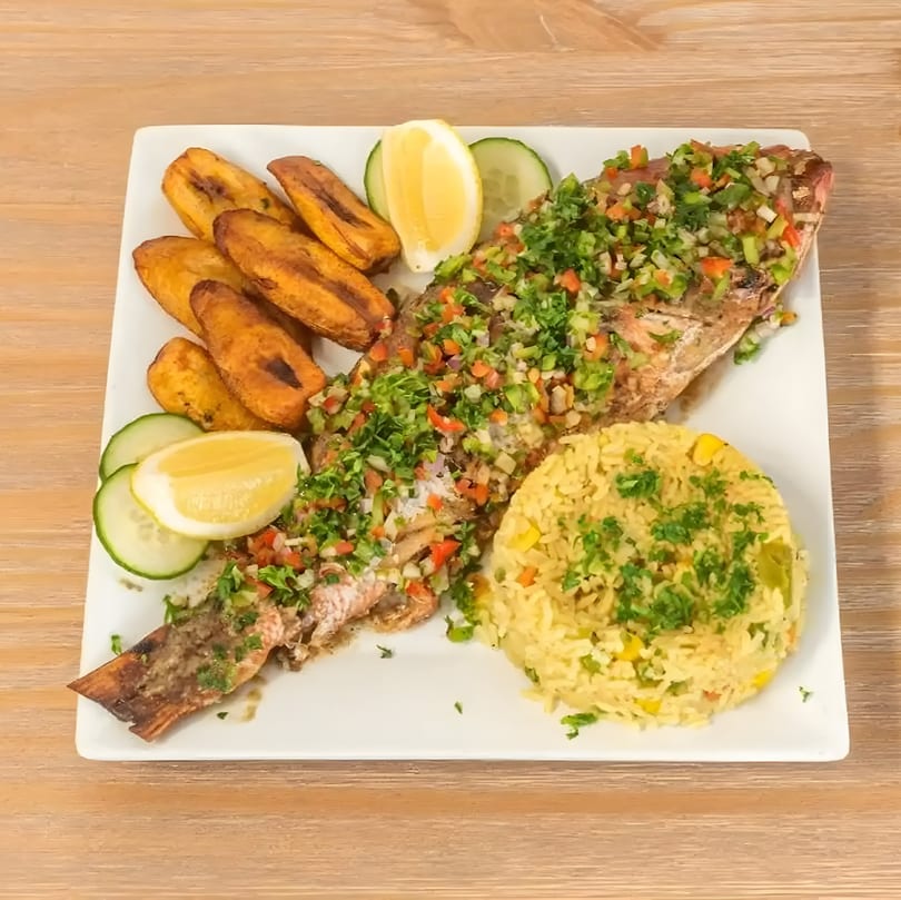 Whole fried fish topped with salsa, served with plantains, rice, lemon, and cucumber slices.