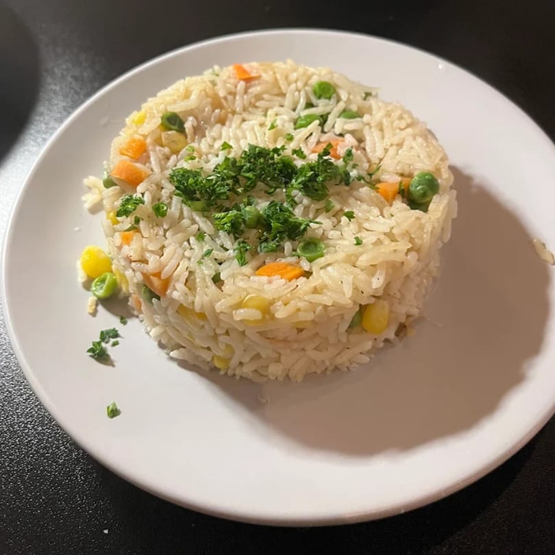 A white plate holds a mound of white rice mixed with peas, carrots, and corn, garnished with parsley.