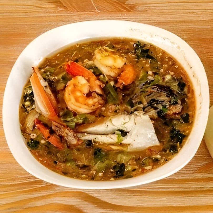 A white bowl of African okra soup with shrimp and crab on a wooden table.