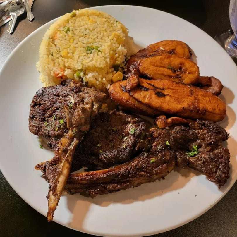 African dish with grilled lamb chops, yellow rice, and fried plantains on a white plate.