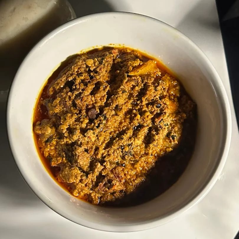 A close-up of a white bowl filled with a rich, textured African stew, possibly Egusi soup.